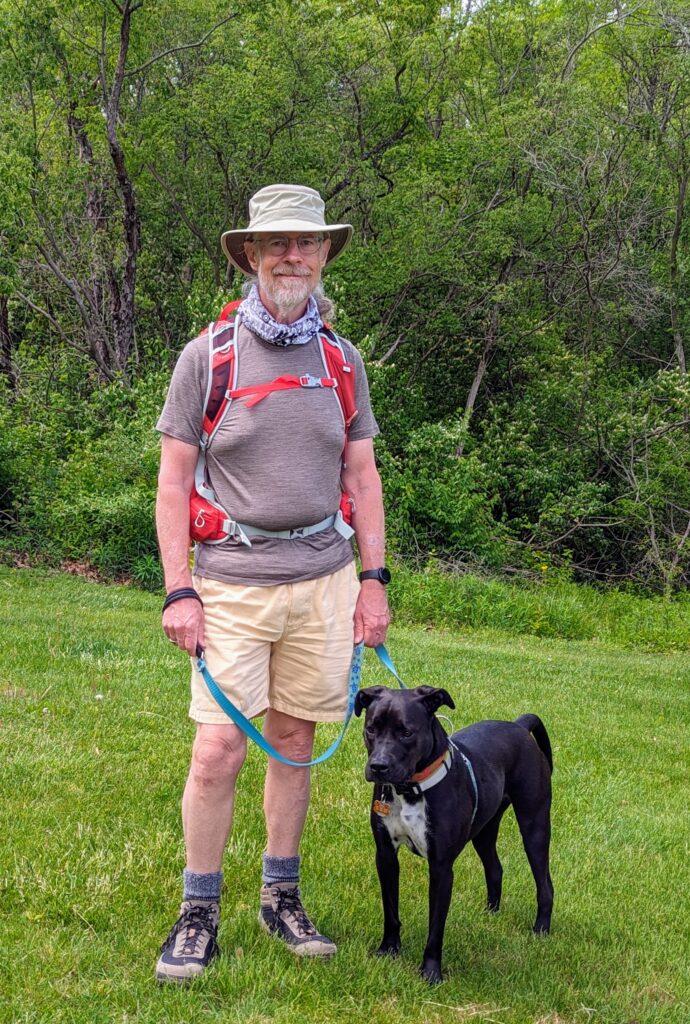 A man in a Tilley hat and a red backpack sending next to a dog that's black with a white chest