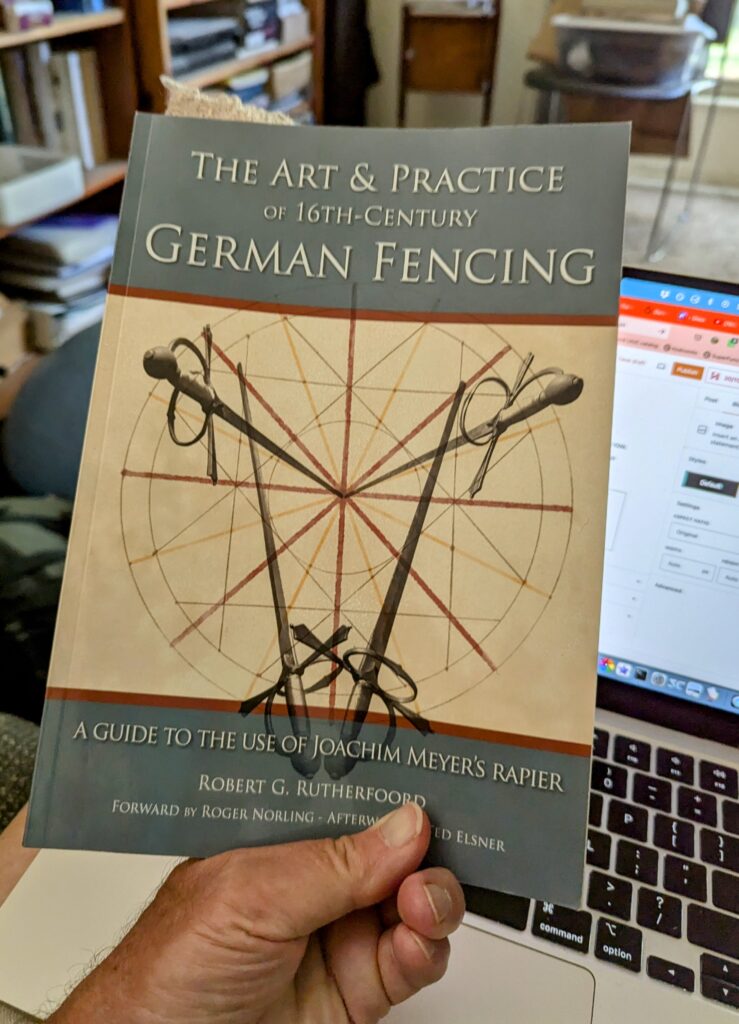 My hand holding a copy of Robert G. Rutherfoord's The Art & Practice of 16th Century German Fencing in front of my laptop