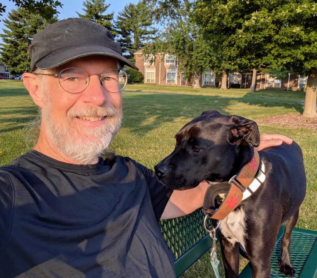 A selfie of me sitting on a park bench with a black dog standing on the bench next to me