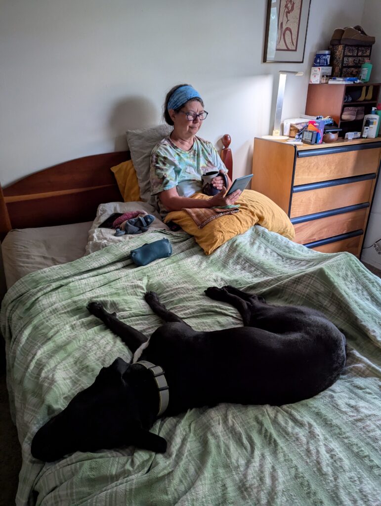 A woman in bed with a big mug of coffee and a kindle, with a dog sprawled across the bed in the foreground