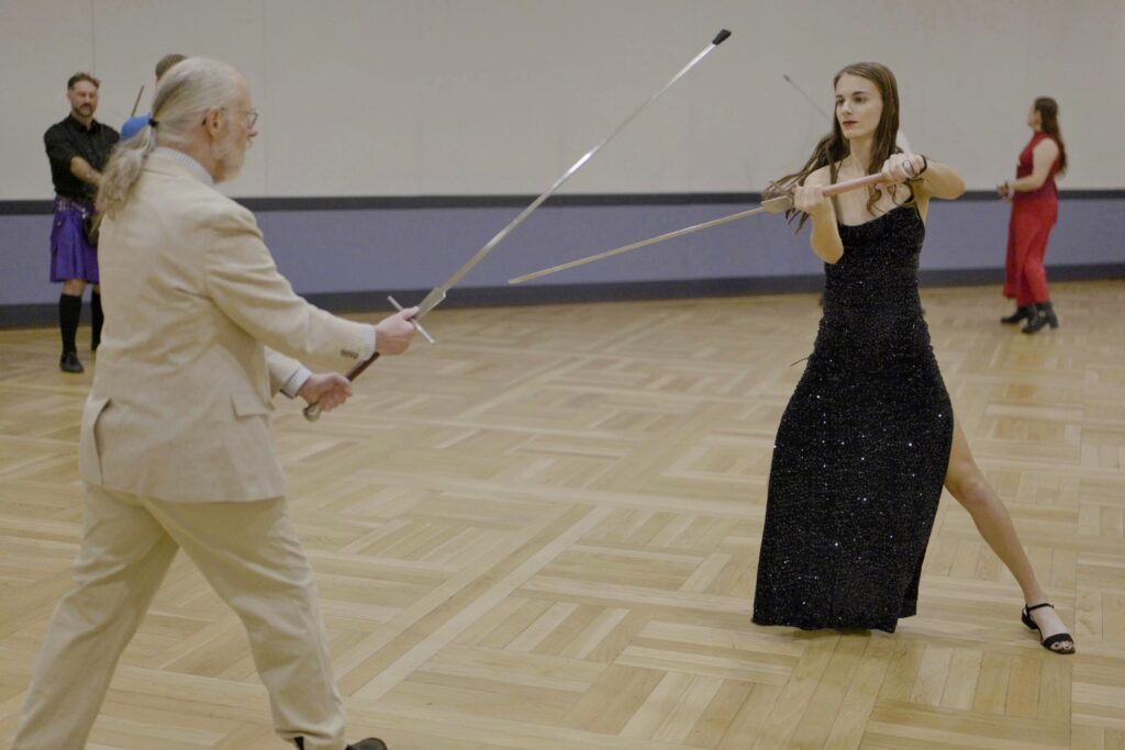 Two people with swords, one wearing a sport coat, the other wearing a fabulous gown, with other people in fancy dress in the background