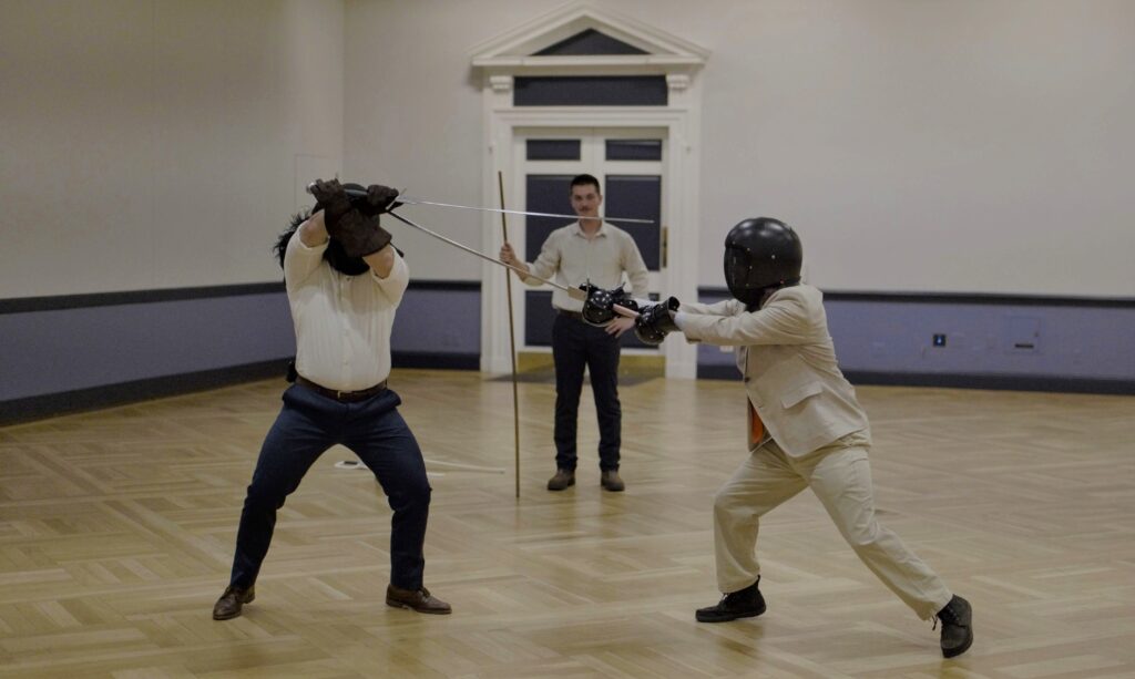 Two fencers in dress clothes with longswords and a judge behind them with a staff